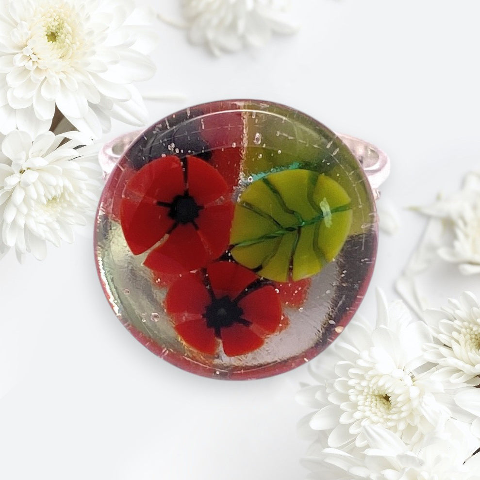 The Calon Glass Poppy Ring (CGR8) is a round handmade glass ring with two red poppy flowers and green leaves inside, in the style of millefiori flower artistry—perfect for nature lovers seeking a unique floral accessory.