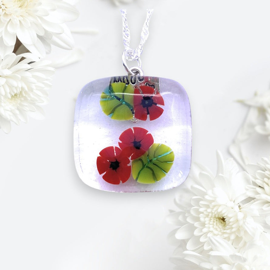 The Calon Glass Poppy Pendant (CGP29) is a handmade glass flower pendant featuring red and green poppy flowers, suspended on a silver chain and displayed among white chrysanthemums.