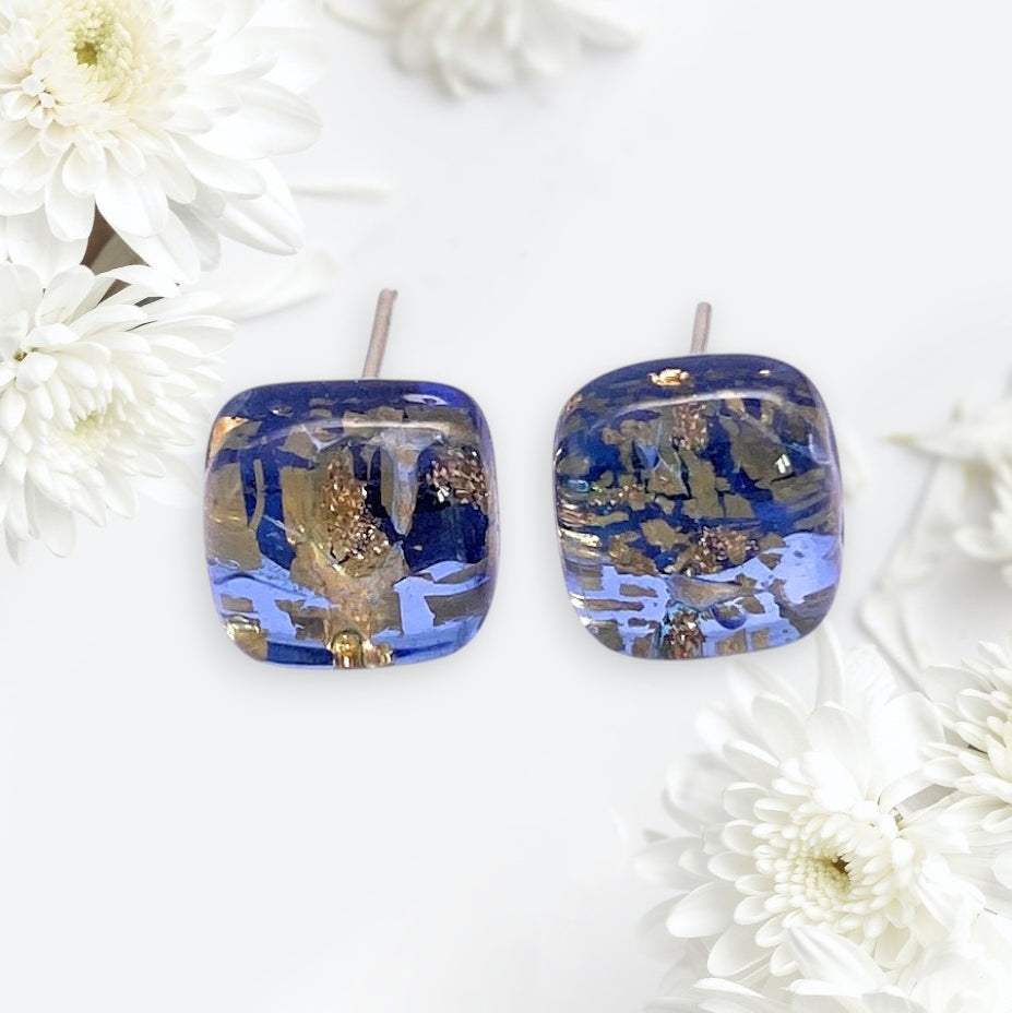 Murano Passion’s Murano Glass Earrings - Blue/Gold (MGE5), dainty 8mm squares with embedded gold flakes, are displayed on a light background amid white chrysanthemum flowers, reflecting Venetian artistry.