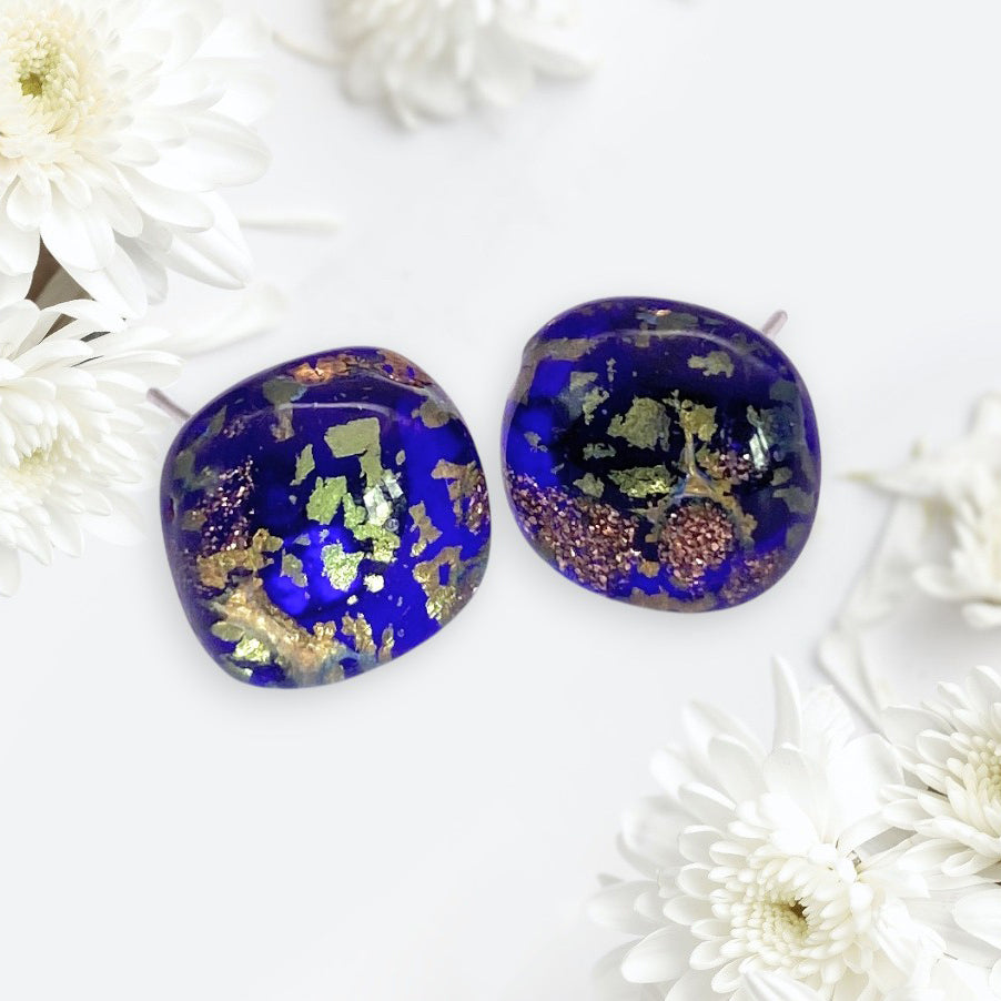 The Murano Passion Murano Glass Earrings (Blue/Gold, 1cm, MGE16) feature round deep blue resin with gold and copper flakes, reminiscent of Handmade Murano Glass, displayed among white chrysanthemum flowers.