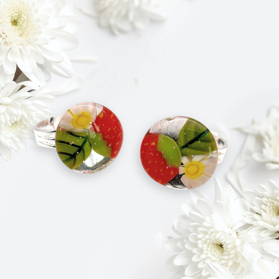 A pair of stunning handmade glass rings, featuring Millefiori floral designs in vibrant red, green, and yellow tones crafted by Calon Glass, is elegantly placed among white chrysanthemums against an immaculate white backdrop.
