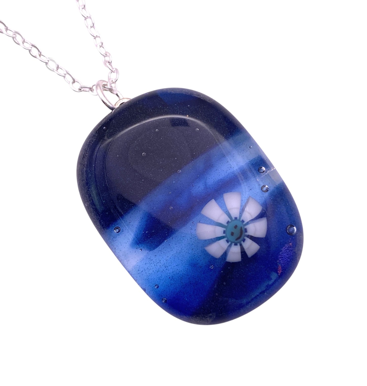 A detailed view of the Calon Glass Smiley Face Millefiori Flower Pendant reveals a blue and black gradient with a white flower, expertly crafted from handmade glass, and elegantly hung on a silver chain. The pendant measures 3cm x 2cm.