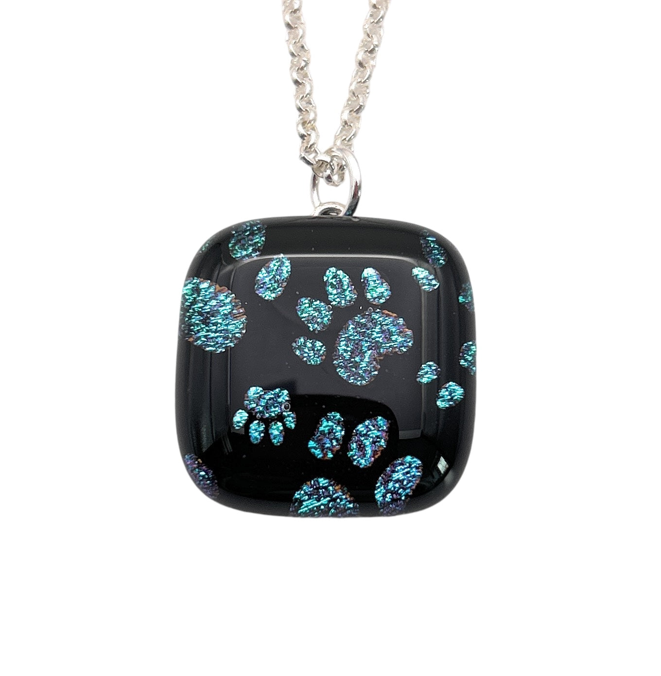 Introducing a distinctive creation from Calon Glass: the Handmade Dichroic Glass 'Paw Print' Pendant, featuring striking blue and teal paw prints set against a black square background. Measuring 2cm by 2cm, this exquisite piece hangs gracefully from a sleek silver chain.