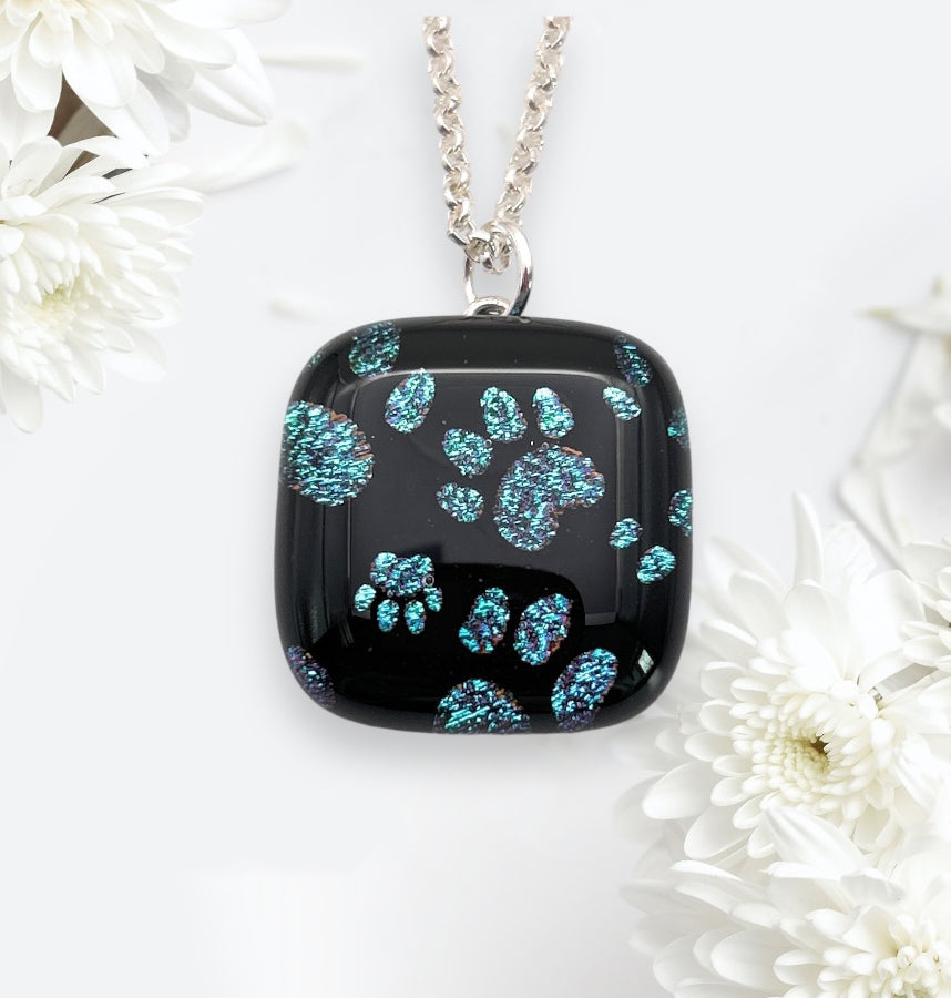 Featuring a 2cm x 2cm square pendant from Calon Glass, this exquisite piece is crafted from handmade dichroic glass and embellished with shimmering aqua paw prints. It hangs gracefully on a silver chain, elegantly framed by white chrysanthemum flowers against a delicate backdrop.