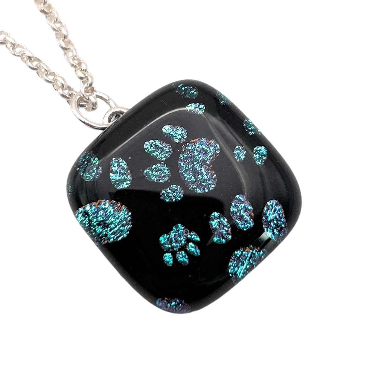 Calon Glass presents a silver-plated necklace featuring the Handmade Dichroic Glass 'Paw Print' Pendant, measuring 2cm by 2cm. The pendant is black with iridescent blue and purple paw prints that shimmer beautifully. Its square shape with rounded edges and glossy finish amplifies the sparkle of the vibrant paw designs.