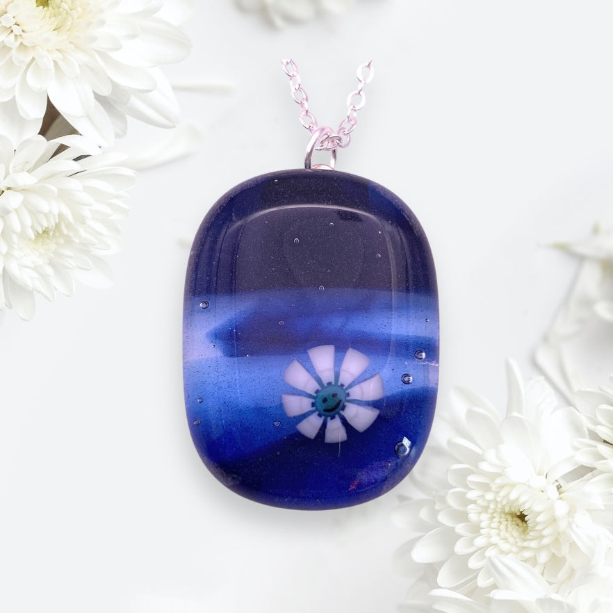 The Calon Glass Smiley Face Millefiori Flower Pendant features a handmade 3cm x 2cm blue glass charm on a silver chain. Surrounded by white chrysanthemums, it creates an elegant and artistic look.