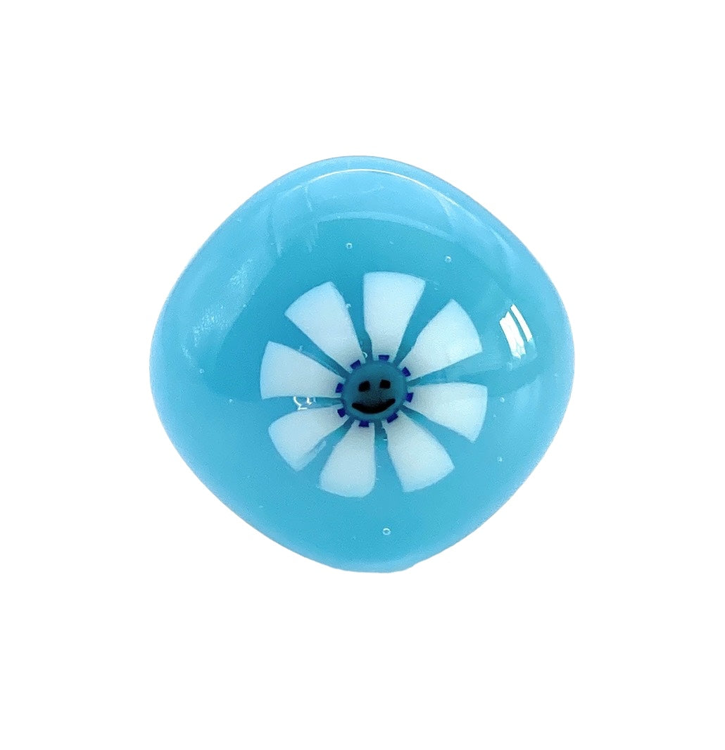This handmade glass ring by Calon Glass features a pod-shaped blue design with a glossy, reflective surface reminiscent of exquisite dichroic glass artistry. At its center is a white circular pattern resembling Millefiori flower petals or fan blades. The ring has a diameter of 1.7cm and is named "Smiley Face Millefiori Flower Glass Ring.