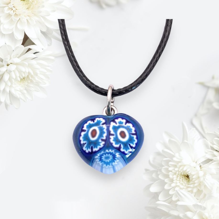 The Murano Passion's 1.4cm Murano Heart Pendant (MHT1) features intricate blue and white floral designs on a striking heart-shaped glass piece, elegantly suspended on a black cord, complemented by white chrysanthemums against a soft background.