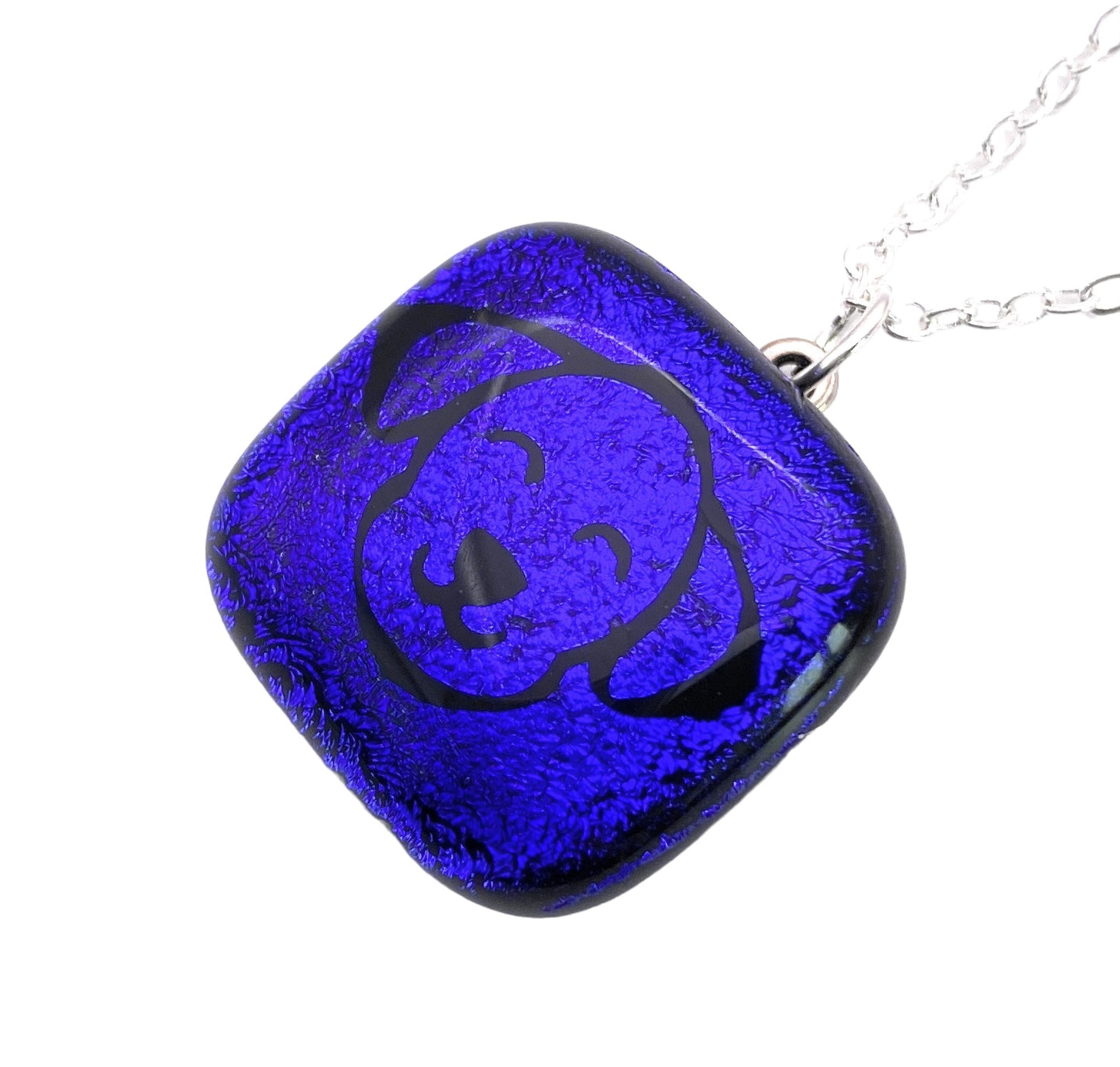 A blue Handmade Dichroic Glass Pendant from Calon Glass, measuring 2cm x 2cm, features a stylized dog face design and hangs from a silver chain. Crafted from kiln-fused glass pieces, it boasts a textured, glossy finish.