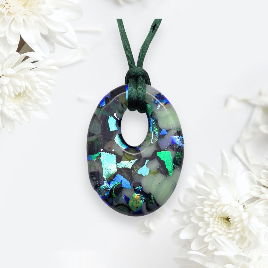 A shimmering oval pendant featuring green, blue, and black mosaic patterns crafted from handmade dichroic glass hangs on a dark green cord. It's from Calon Glass and measures 4cm x 3cm (Product ID: CGP34), set against a light background with white flowers surrounding it.