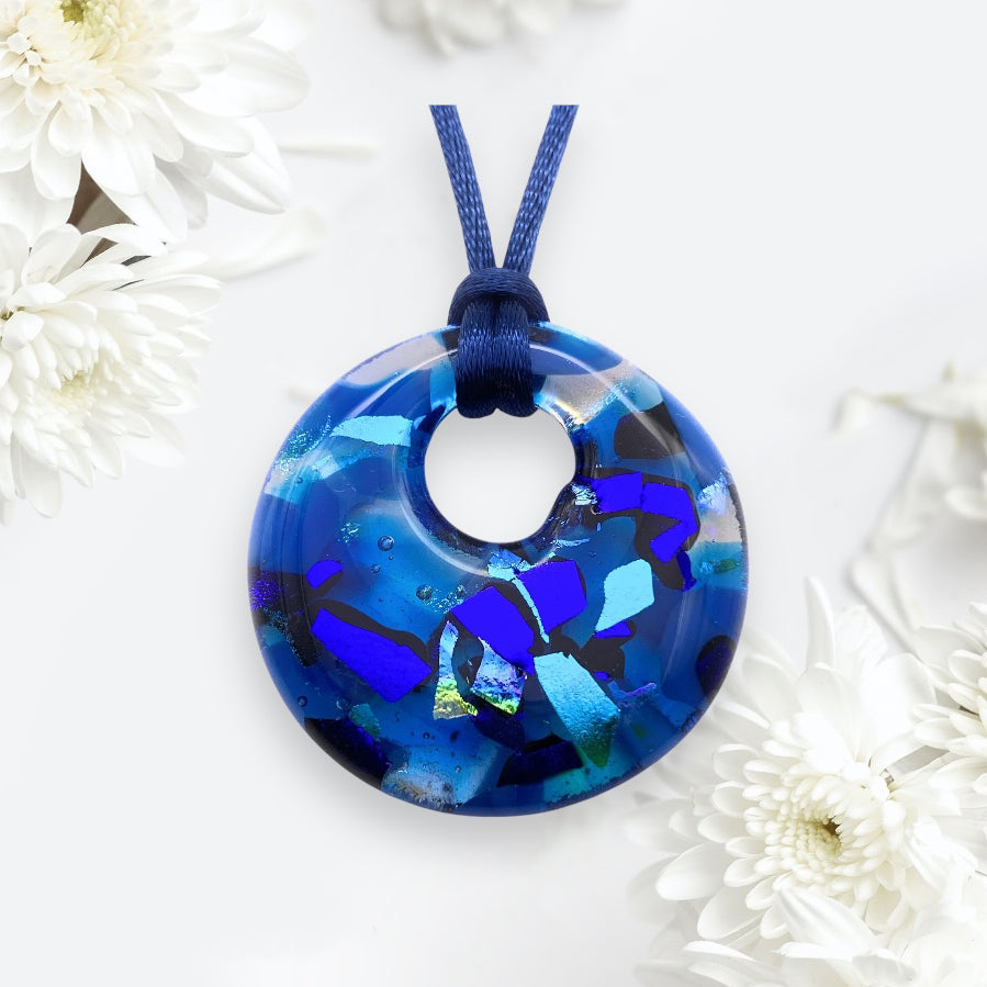 A round, kiln-fused blue dichroic glass pendant with abstract patterns in lighter and darker shades is hung on a blue cord. The Handmade Dichroic Glass Pendant by Calon Glass, model CGP33, is beautifully surrounded by white flowers on a light background.