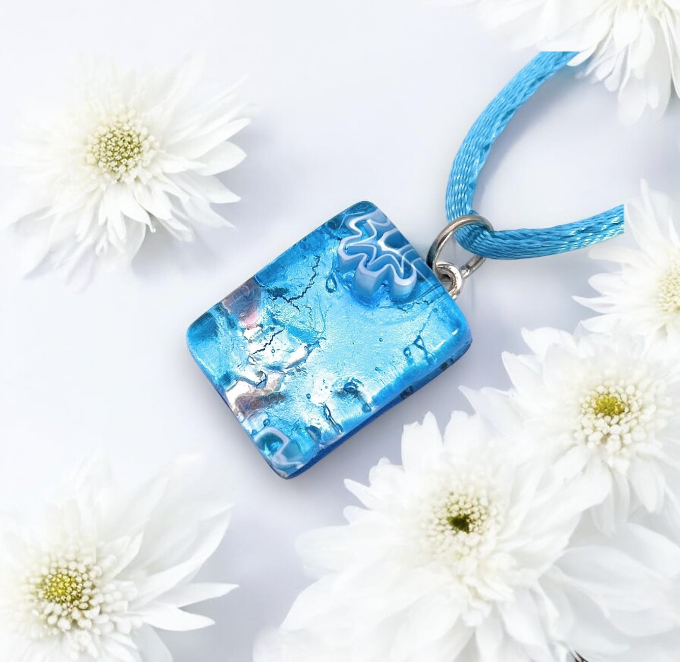 The Murano Passion MGPP18 is a 2cm x 1.5cm rectangular pendant featuring marbled blue and white patterns on a blue cord, surrounded by delicate white flowers on a light background. Crafted with silver leaf, this authentic Murano Glass Jewelry includes a Certificate of Authenticity.