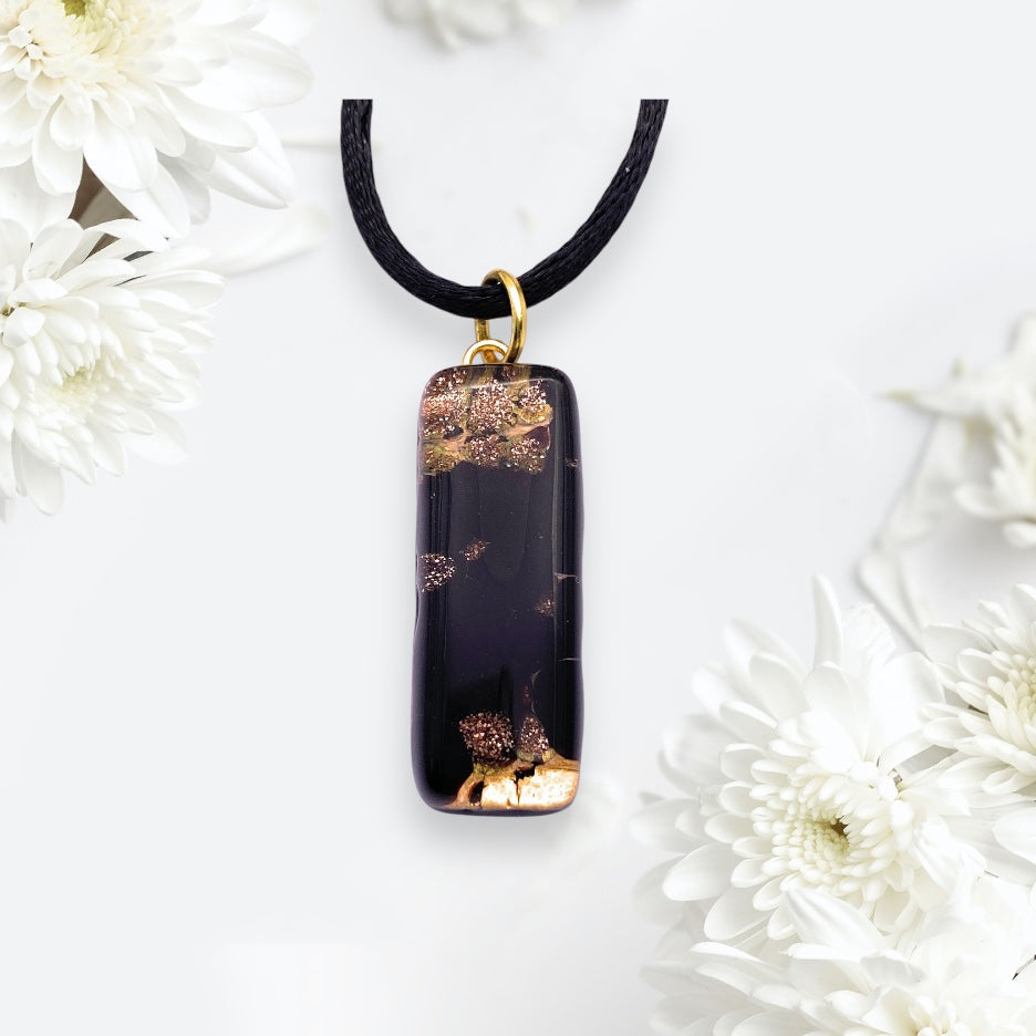 The Murano Passion MGPL5 pendant, featuring rounded edges and luxurious gold leaf on black Murano glass, hangs from a black cord. This exquisite 3cm x 1cm handmade piece is beautifully framed by white flowers against a soft white background.