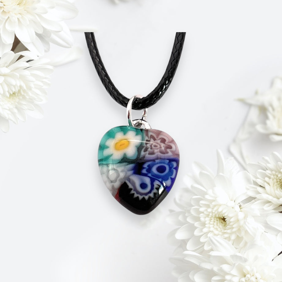 The Murano Passion Murano Glass Pendant, a 1.4cm heart-shaped piece with colorful floral designs, hangs from a black braided cord. White flowers accentuate its light background, and it includes a Certificate of Authenticity for assurance.