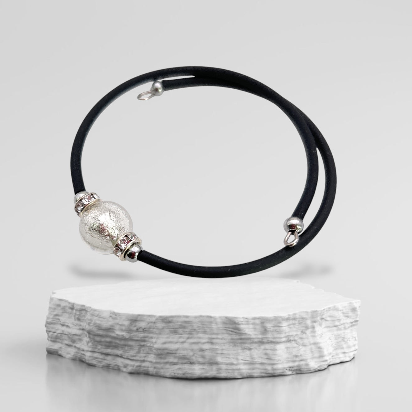 A Murano Passion Murano Glass Bead Bracelet, a handmade Memory Wire piece featuring a textured silver Murano glass bead and smaller metallic accents, is elegantly displayed on a white, textured platform against a plain gray background.