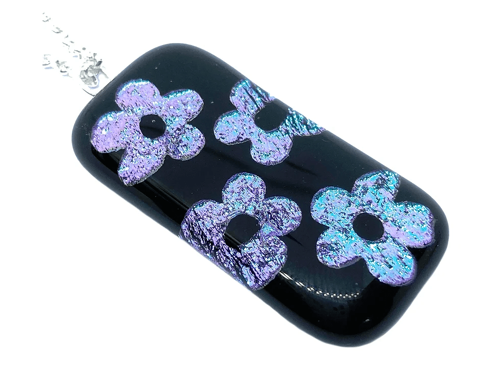 The Calon Glass Handmade Dichroic Glass Pendant (4cm x 2cm) features four iridescent purple flower designs on a rectangular black base, finished with a silver chain.
