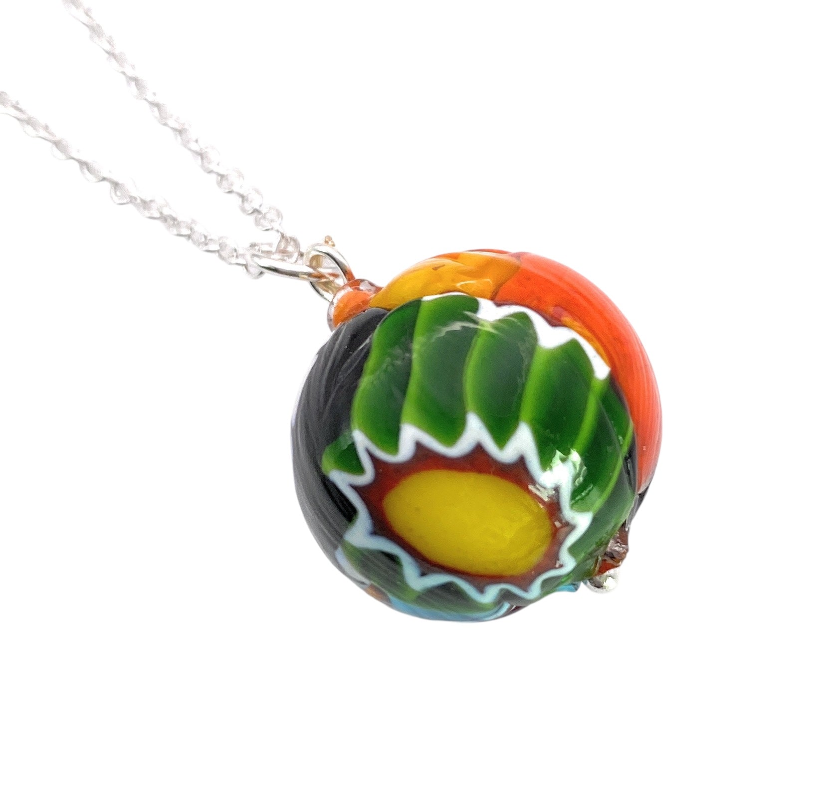 This eye-catching piece from Murano Passion, the Murano Glass Multi Coloured Millefiori Ball Pendant (MGB1), features a stunning array of vibrant colors including green, yellow, and orange. The pendant displays swirling patterns with a central yellow circle outlined by white zigzag designs, all suspended from a sleek silver chain. A hint of Aventurine glass enhances its sparkling charm.