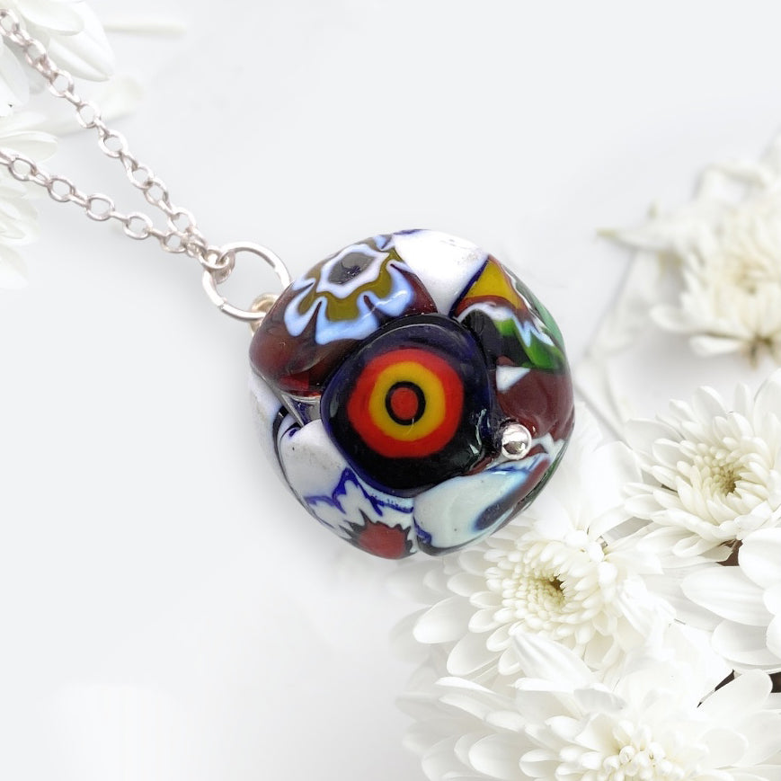 Introducing the Murano Passion's Murano Glass Multi Coloured Millefiori Ball Pendant (MGB2), this pendant artfully showcases abstract floral designs in vivid hues of red, yellow, and blue. Suspended beautifully on a silver chain, it is complemented by a soft white floral background and includes a Certificate of Authenticity.