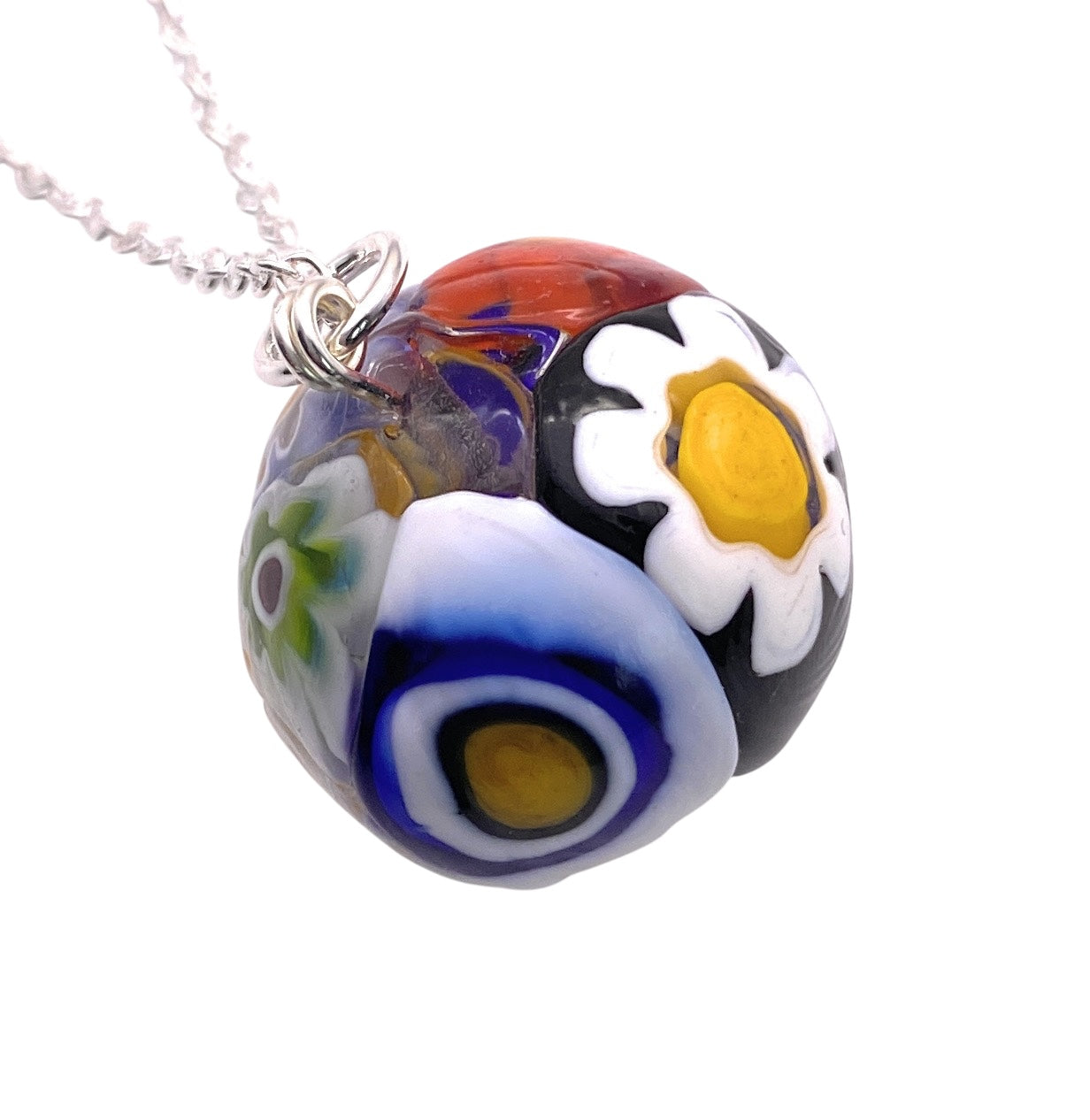 The Murano Passion's Millefiori Ball Pendant (MGB4) is a vibrant piece of jewelry crafted with aventurine glass. It features intricate floral designs, including daisies and abstract patterns, showcased in a colorful palette of red, blue, black, and white. Hanging from a silver chain, this Murano glass pendant is truly captivating.