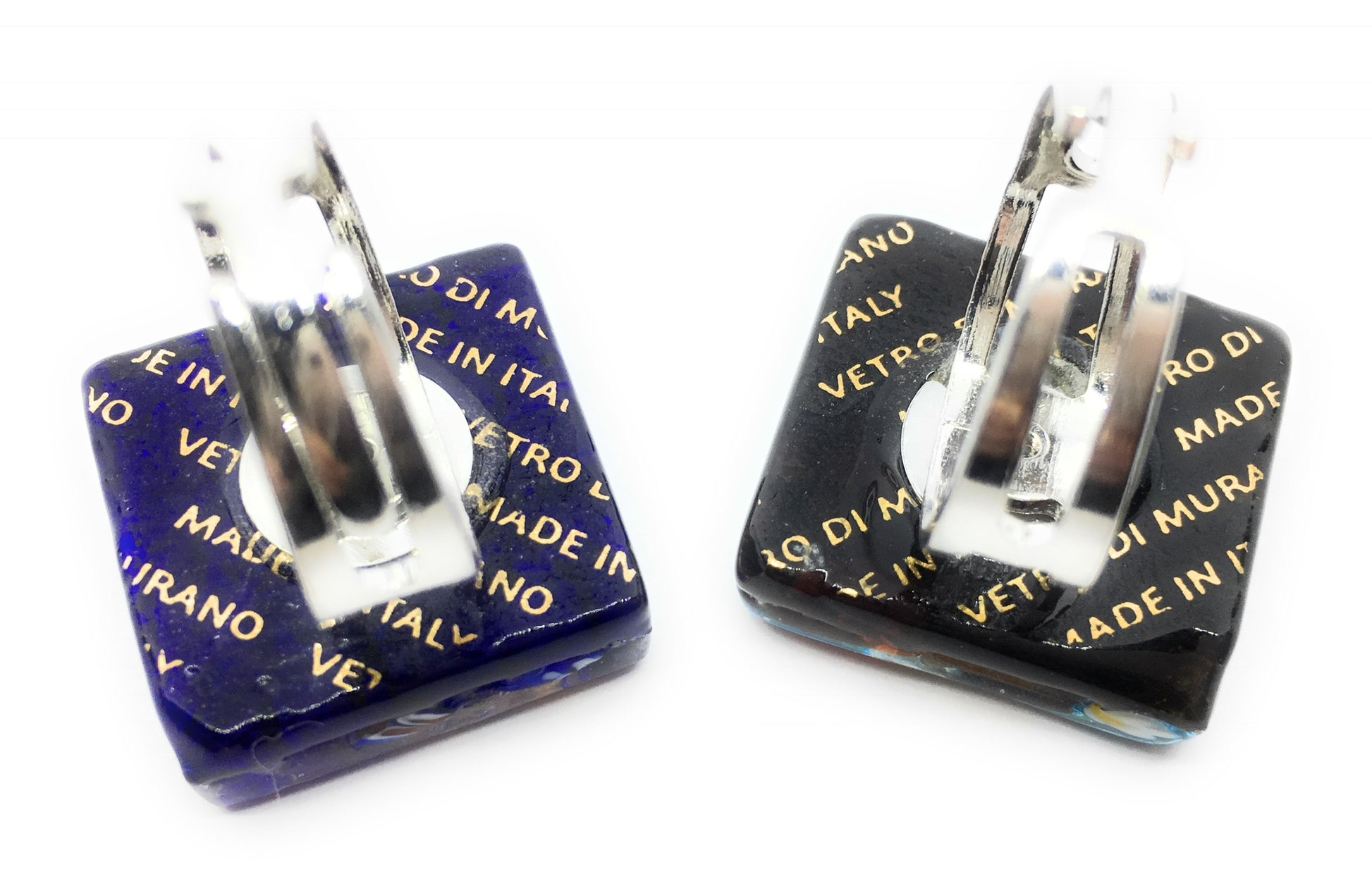 Two square glass cufflinks featuring metallic text are positioned next to each other. The left cufflink showcases a dark blue hue with gold writing, while the right is black—crafted entirely from authentic Murano Glass. Each item is elegantly finished with silver clasps, bearing the inscriptions "Made in Italy" and "Vetro di Murano.