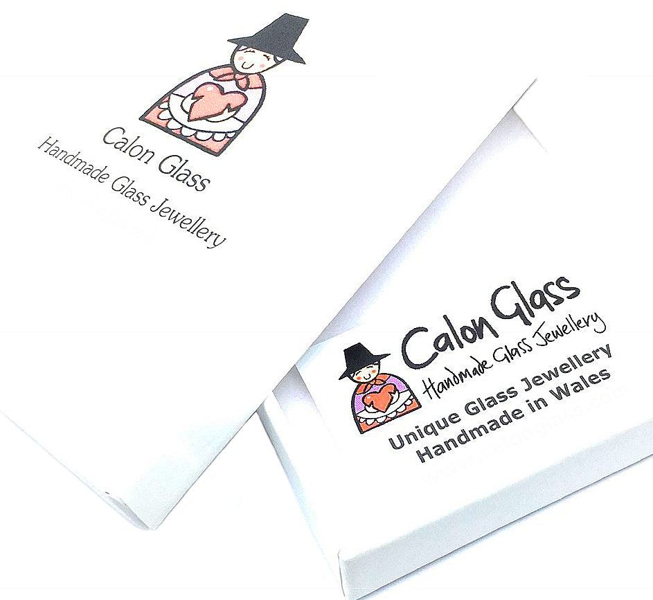 Two white packaging boxes bear the "Calon Glass" logo, known for unique handmade jewellery crafted with dichroic glass. The text reads, "Unique Glass Jewellery Handmade in Wales." An illustration shows a person in traditional Welsh attire with a heart.