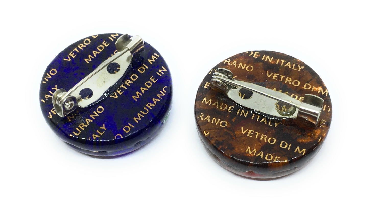 Two Murano Passion circular brooches with metal fasteners on the back beautifully showcase Venetian artistic heritage. The left brooch features a dark blue design, while the right one is brown, both embellished with "MADE IN ITALY" and "VETRO DI MURANO" in gold lettering. Each Murano Glass Brooch, with a silver leaf base and a 2.4cm diameter, exudes handmade elegance.