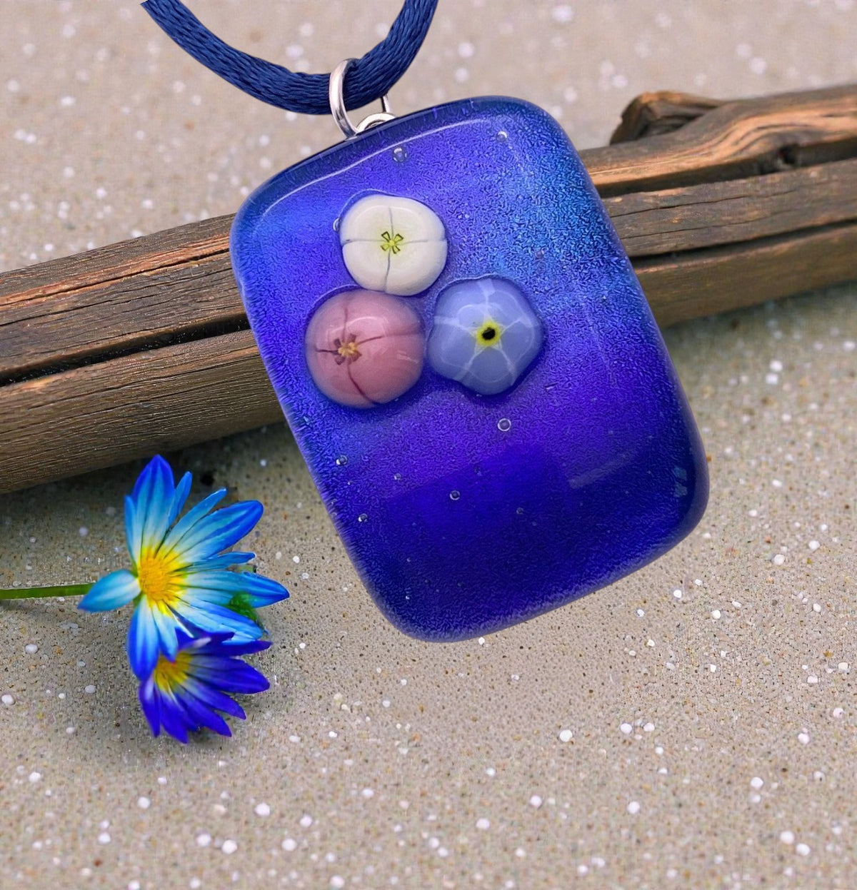Handmade Dichroic Glass Pendant, Shiny Blue Dichroic Glass with Millefiori Flowers (CGP21) featuring a rectangular blue pendant with three small millefiori flower embellishments, hanging on a dark blue cord.