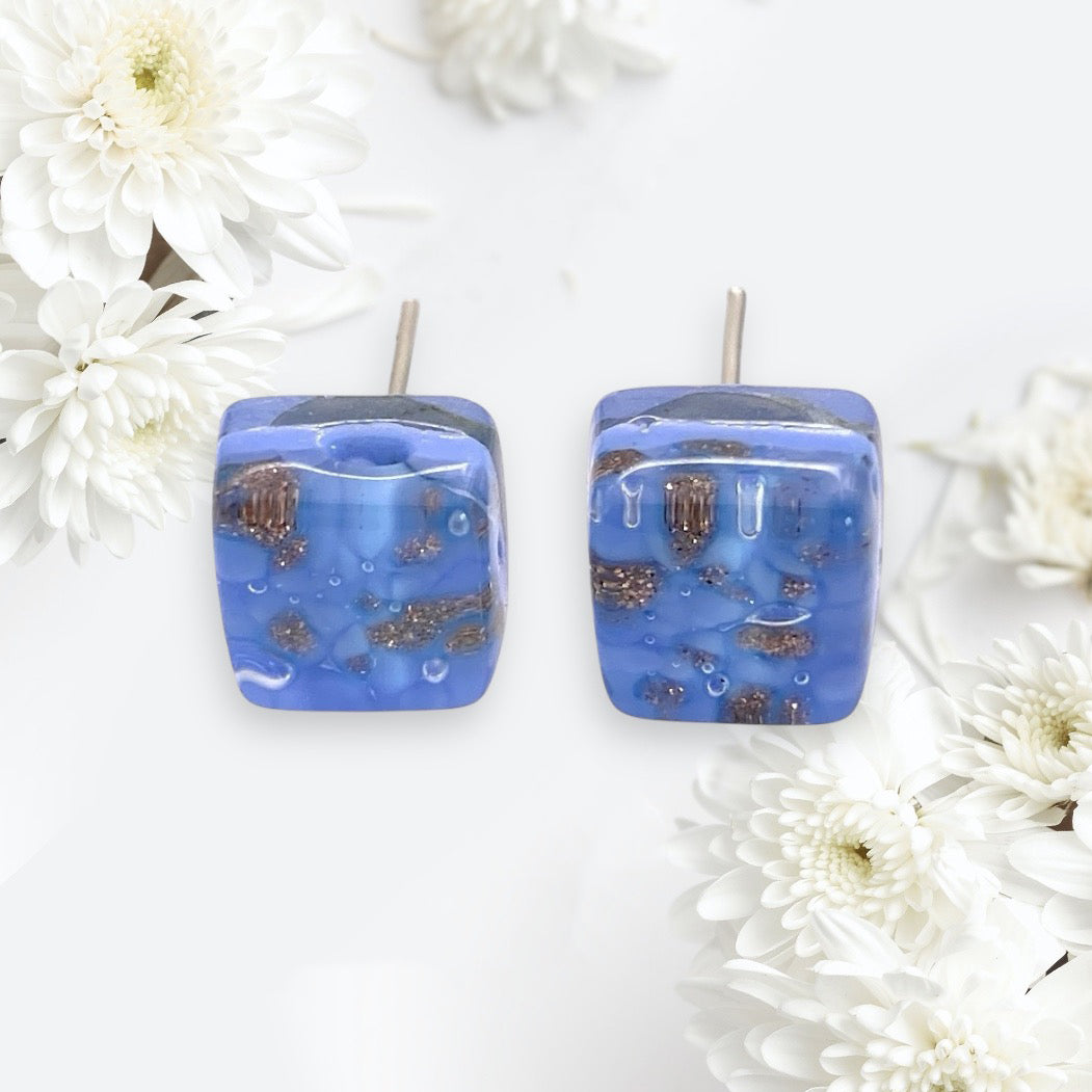 Dainty, square resin earrings in a striking blue feature embedded brown accents and shine brilliantly against the backdrop of white flowers. These post-style Murano Passion treasures boast a shiny finish that echoes the elegance of Murano Glass Earrings. Meticulously crafted, each pair is celebrated as an 8mm diameter handmade masterpiece from their exquisite collection (MGE16).