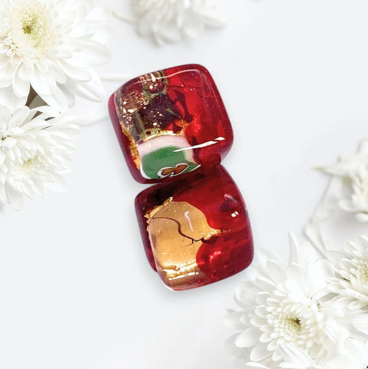 Two glossy red glass earringse with gold and green swirls rest on a white surface among soft chrysanthemums, echoing the intricate beauty of Murano Passion’s Red/Gold Dainty 8mm Murano Glass Earrings (MGE9).