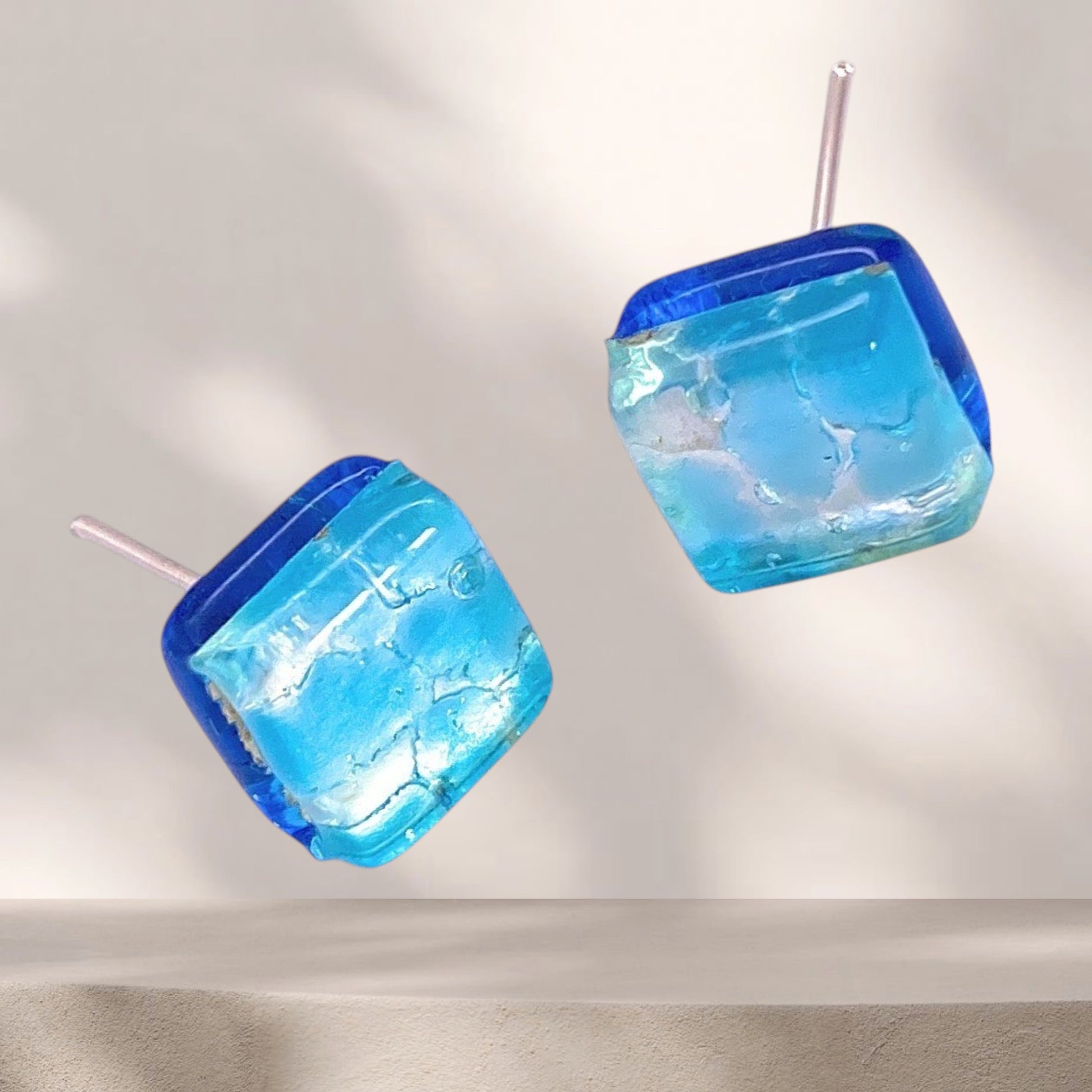 Murano Passion’s Murano Glass Earrings (MGE18) feature two dainty 8mm pale blue squares with an icy, translucent look and silver posts, displayed on a neutral background.