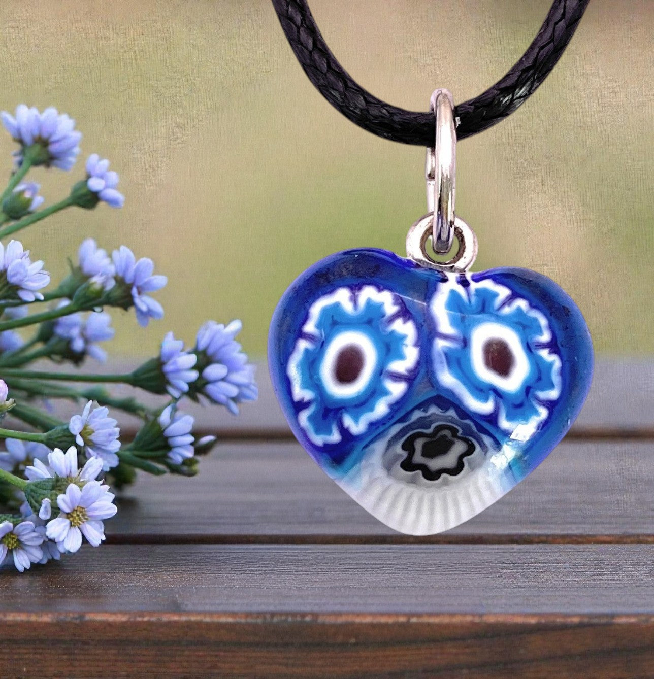 A delicate, heart-shaped blue Murano Glass Pendant by Murano Passion (Murano Heart Pendant - Tiny 1.4cm, MHT6), reminiscent of exquisite Venetian craftsmanship, showcases intricate floral patterns with small purple flowers on the left. The pendant hangs gracefully from a black cord and stands out beautifully against a backdrop of blurred greenery.