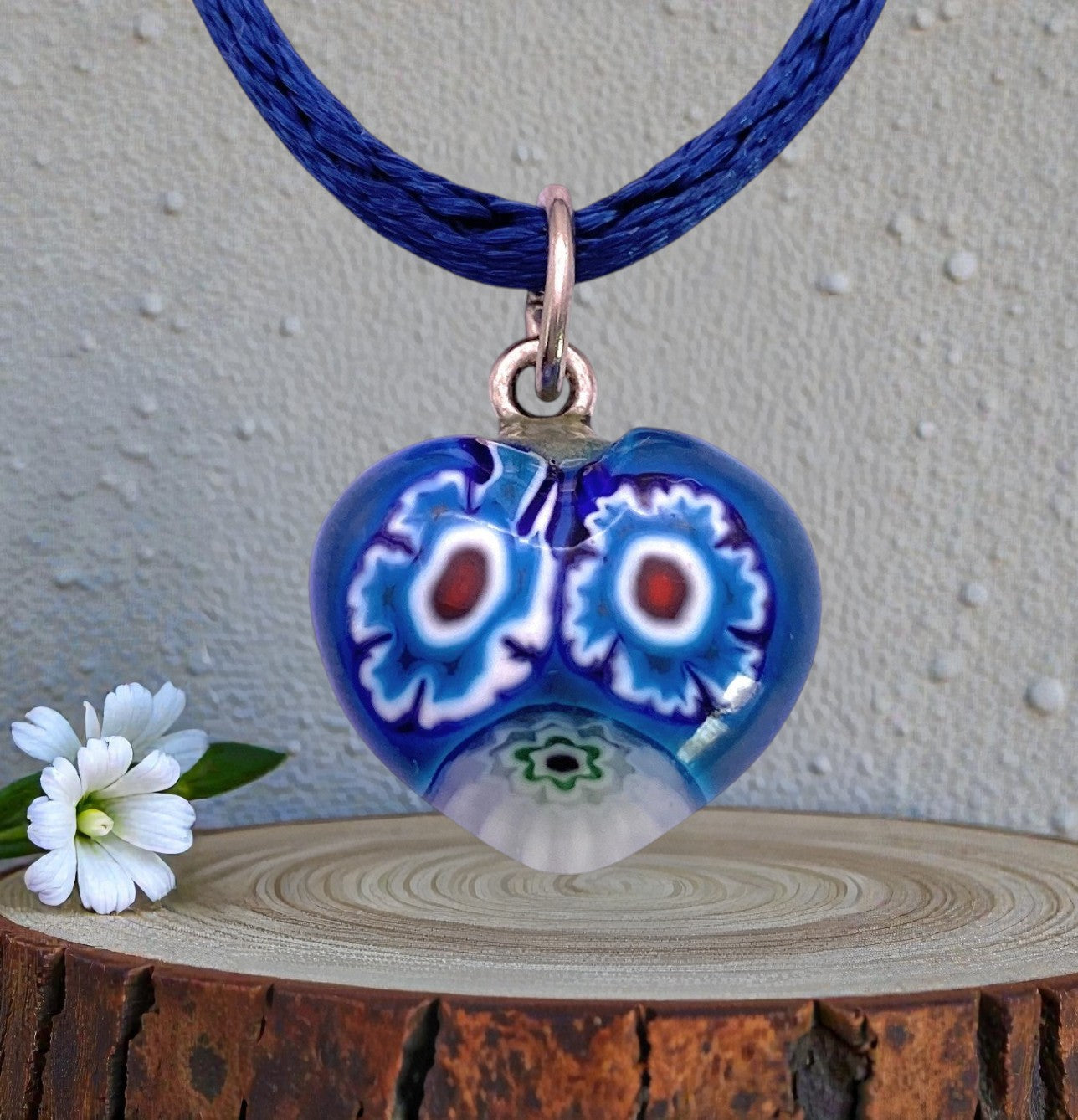 The Murano Passion Murano Heart Pendant (Tiny 1.5cm, MHT3) features colorful floral patterns on blue Murano glass and hangs on a blue cord, displayed beside a small white flower on a wooden surface.
