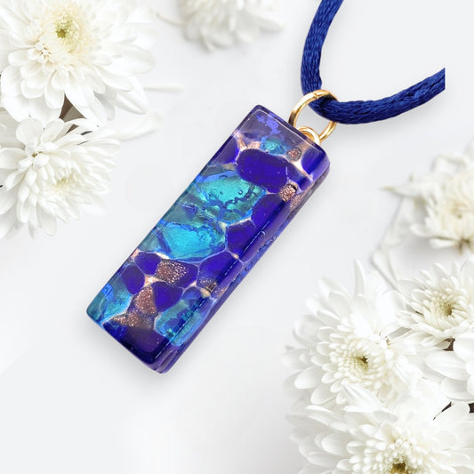 The Murano Passion Murano Glass Pendant with Blue on Gold Leaf (3cm x 1cm, MGPL11) is shown on a dark blue cord among white flowers, highlighting its elegant marbled design.