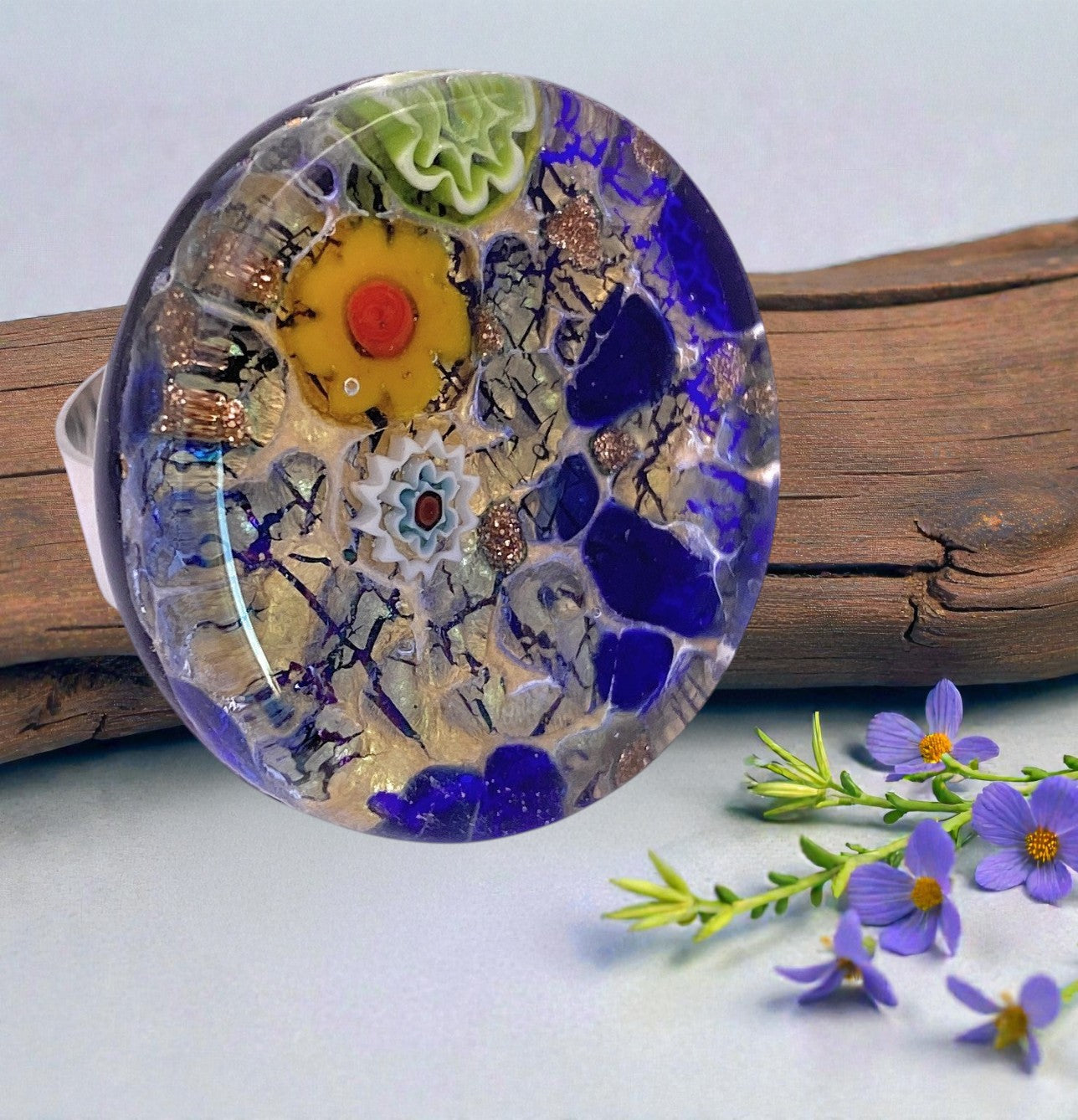 A chunky 3cm Murano Passion glass statement ring (MGR3) with vibrant colors and intricate designs rests on a wood surface, surrounded by small purple flowers and green sprigs.