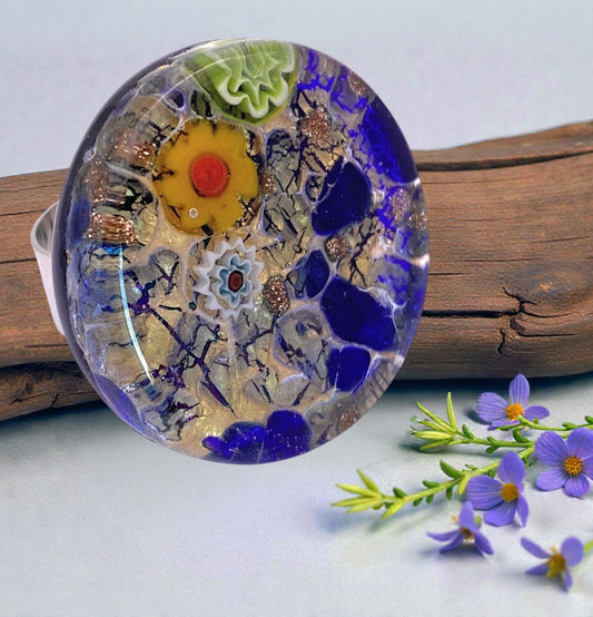 A chunky 3cm Murano Passion glass statement ring (MGR3) with vibrant colors and intricate designs rests on a wood surface, surrounded by small purple flowers and green sprigs.