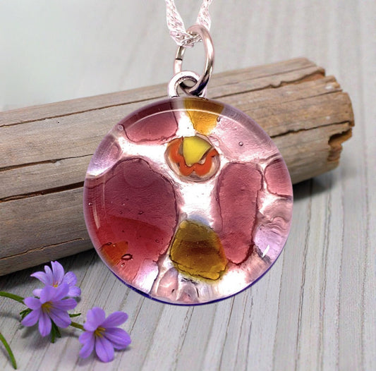 The Murano Passion Murano Glass Round Pendant made with Millefiori (2cm, MGPV1) features vibrant abstract colors on a silver chain, displayed on light wood with purple flowers—Venetian artistry in every detail.