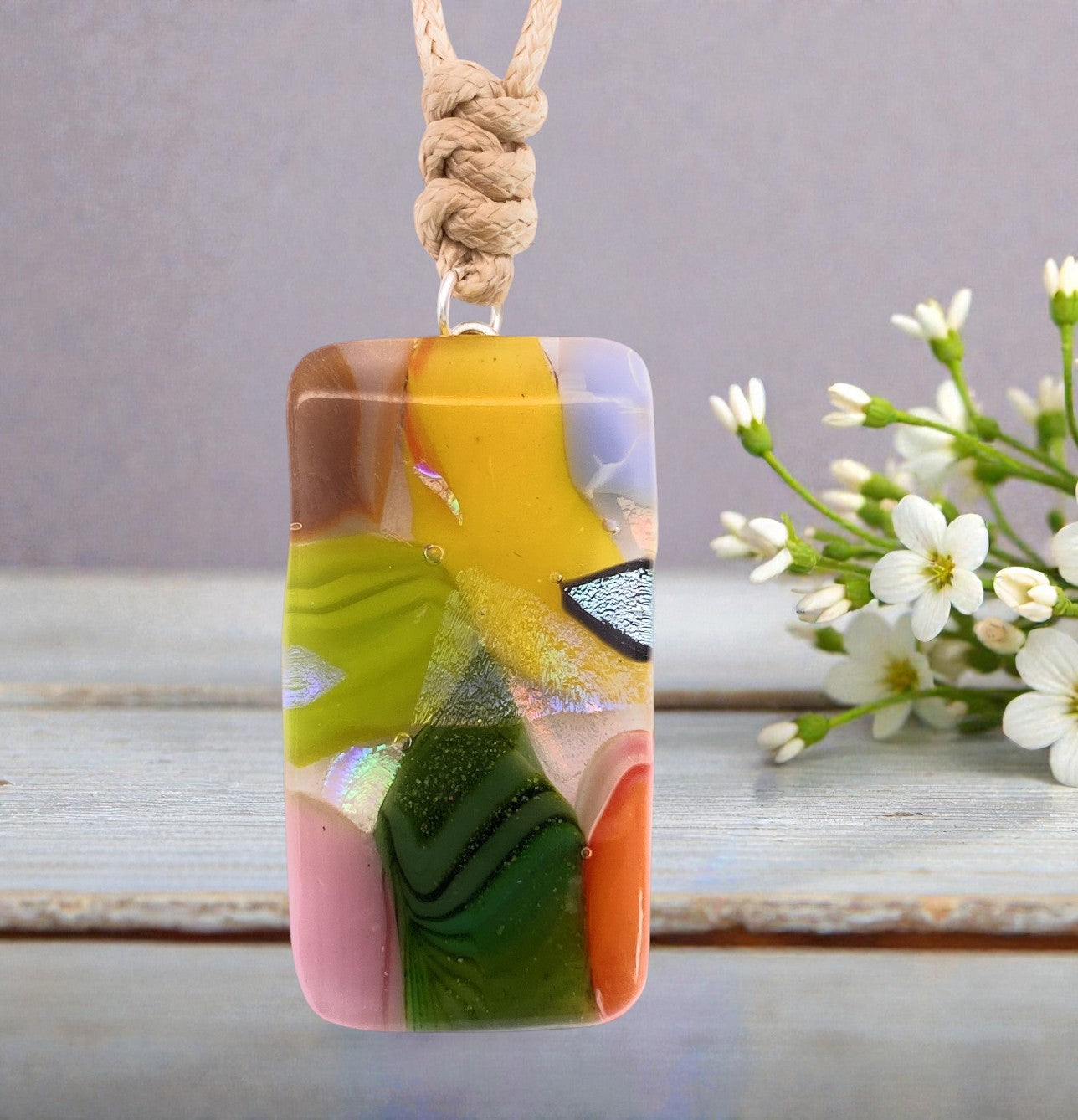 The Handmade Dichroic Glass Mosaic Pendant – Multicolour Fused Glass Statement Necklace (CGP82) by Calon Glass displays vibrant abstract patterns and hangs from a beige cord, set against a backdrop of delicate white flowers and a light wooden surface.