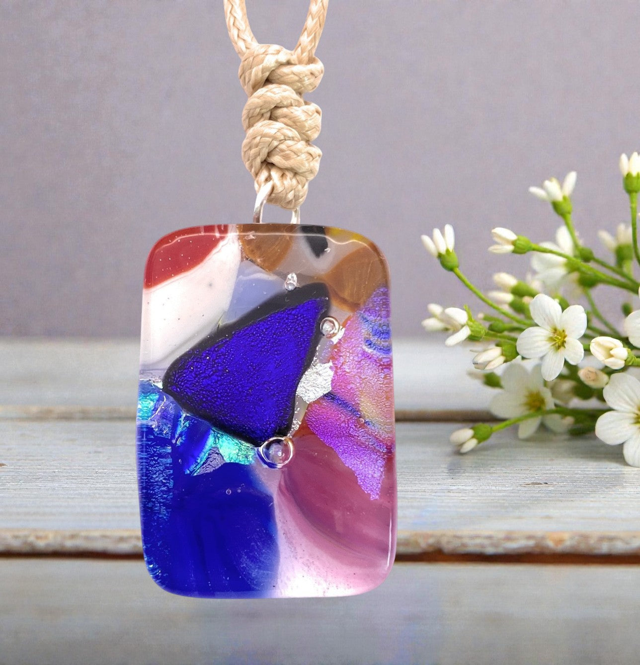 The Calon Glass Handmade Dichroic Glass Mosaic Pendant – Multicolour Fused Glass Statement Necklace (CGP83) features a rectangular design with abstract swirls of blue, purple, pink, white, and gold on a braided cord, beautifully displayed next to small white flowers on a wooden surface.