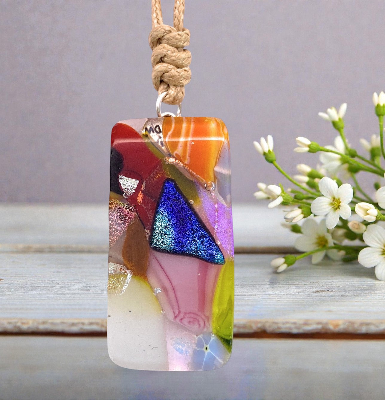 Showcased on a wooden surface with delicate white flowers in the background, the Calon Glass Handmade Dichroic Glass Mosaic Pendant – Multicolour Fused Glass Statement Necklace (CGP84) features abstract shapes and a vivid blue triangle. This colorful, artisan pendant hangs from a beige cord, making it a striking and unique accessory.