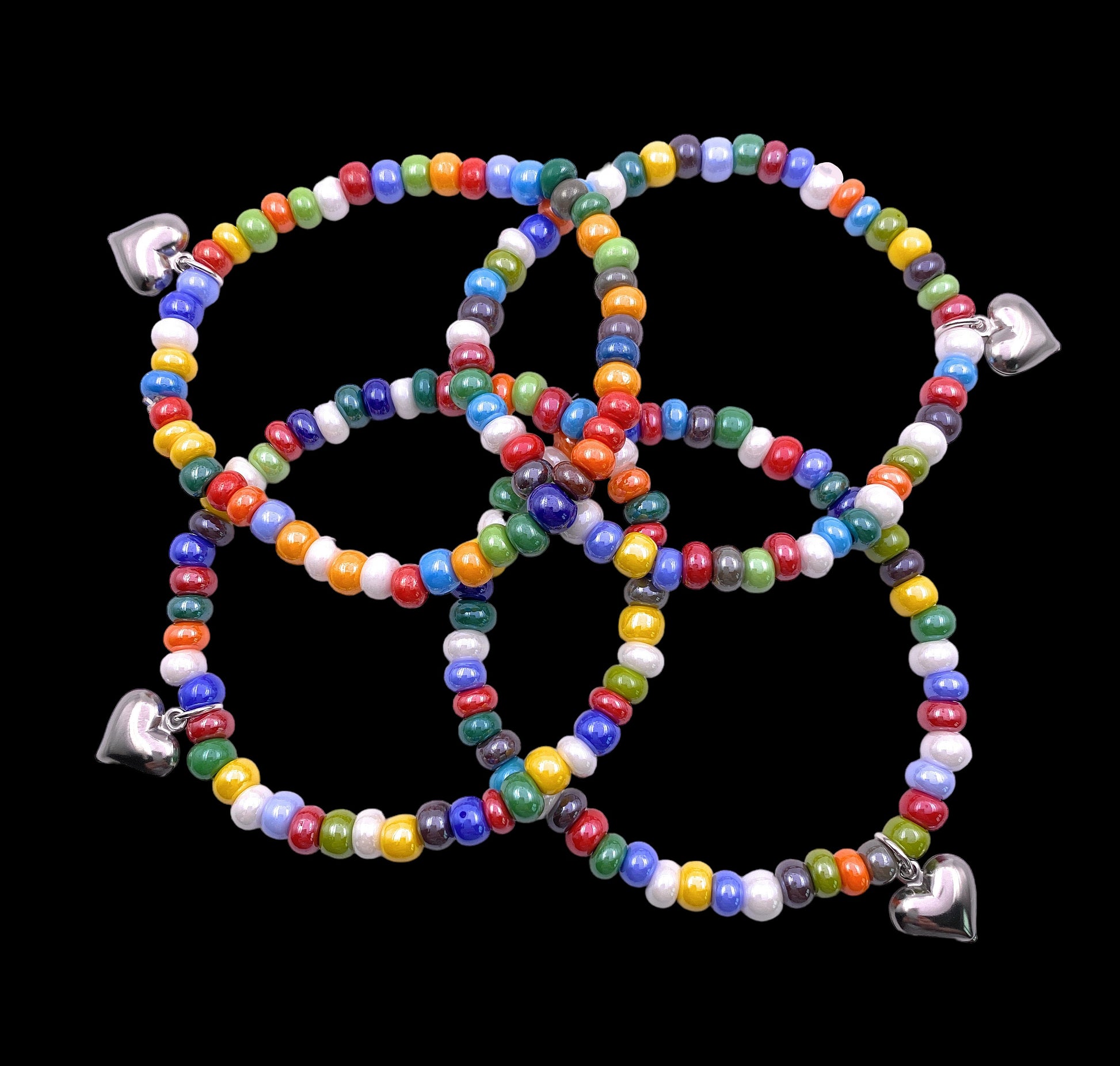 Five vibrant bracelets from Murano Passion, each crafted with 5mm/0.5cm multi-colored Venetian glass beads and adorned with a delicate silver heart charm, are elegantly interlocked on a black background. These handmade Murano Glass Beaded Bracelets (MGBR7) capture the enchanting beauty of artisanal jewelry.