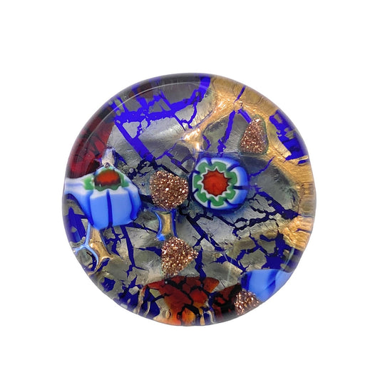 The Murano Passion brooch, named the Murano Glass Brooch with Millefiori on a Gold Leaf Base, showcases a circular abstract design and intricate, colorful patterns. Handmade in Venice, Italy, it features blue, gold, and red designs accented with small shimmering brown details on a white background.