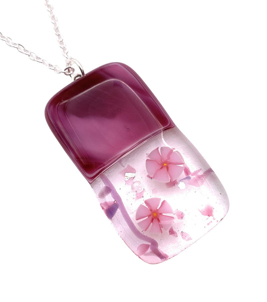 A stunning Handmade Dichroic Glass Pendant by Calon Glass, measuring 4cm x 2cm, showcases a rectangular design with a deep pink top half and clear bottom half. It elegantly features embedded Millefiori flowers and sparkling glitter, all suspended from a silver chain—a true testament to the beauty of handmade glass craftsmanship.