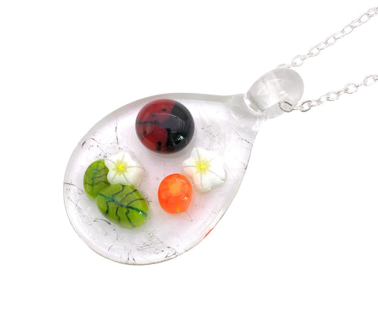 The Calon Glass handmade glass pendant, also known as the Ladybird Pendant, features a teardrop shape adorned with vibrant millefiori designs. It showcases colorful orbs including one red, two green with leaf patterns, and one orange, along with two white flowers inside. This floral pendant hangs elegantly from a silver chain and measures 3cm by 2.5cm.