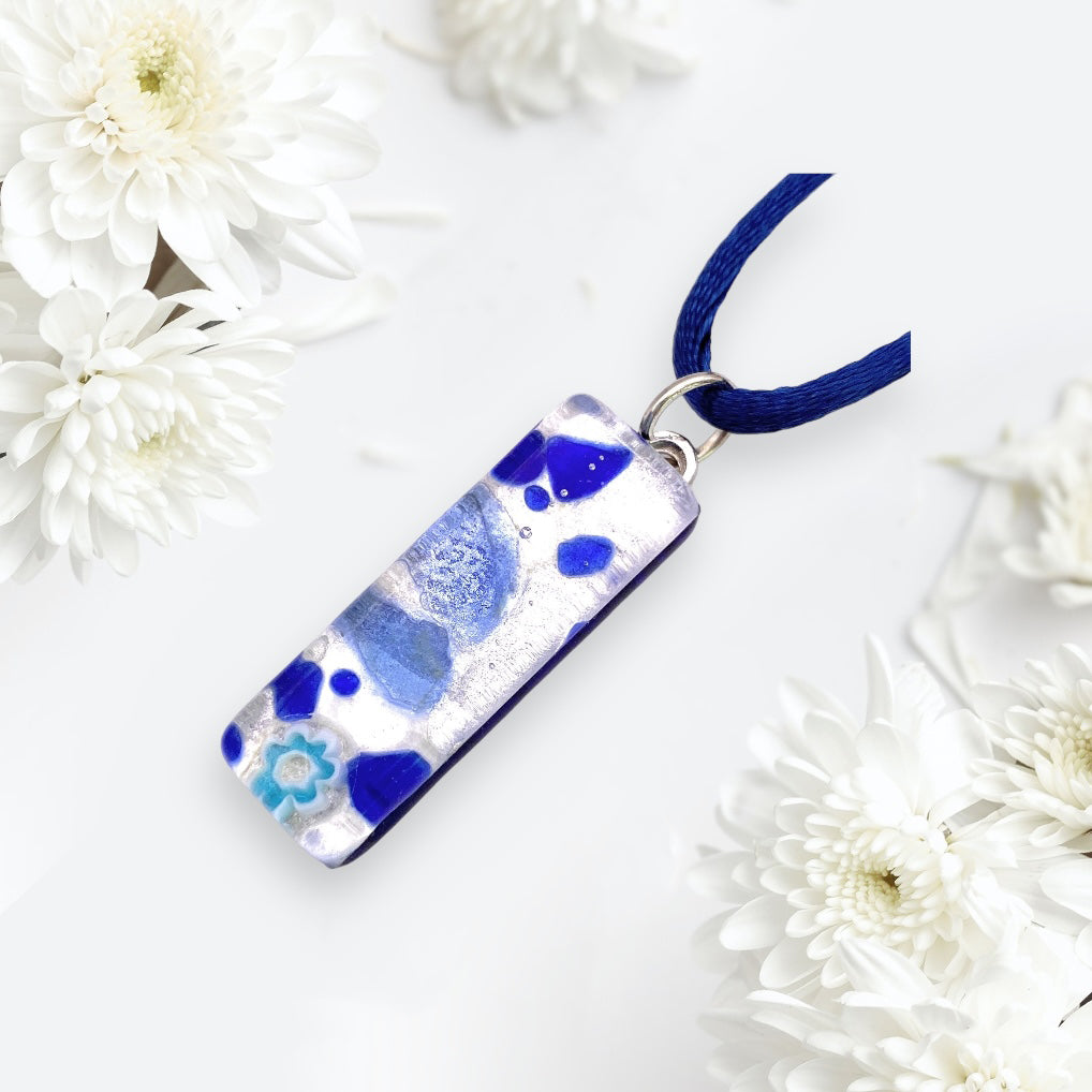 A Murano Passion Murano Glass Pendant on Silver Leaf (3cm x 1cm, MGPL4) with blue and white abstract patterns hangs from a blue cord, set against a backdrop of white flowers.