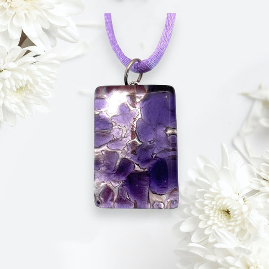 A detailed view showcases a rectangular Murano Passion Glass Pendant, featuring intricate Millefiori Flower patterns on silver leaf, suspended from a purple cord. The pendant is surrounded by white flowers set against a light backdrop, enhancing its soft elegance. Complete with its Certificate of Authenticity, this 3cm x 2cm MGPC2 piece radiates timeless beauty.