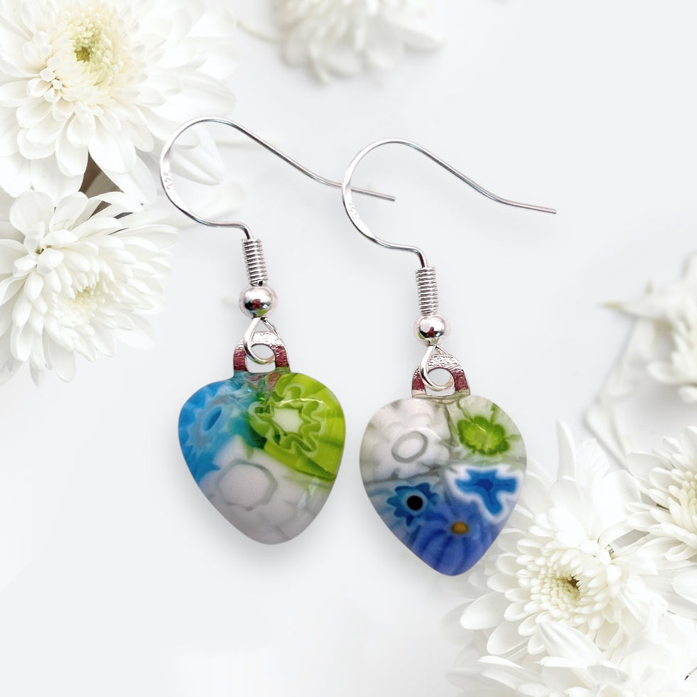 Heart-shaped earrings with a colorful floral pattern hang against a soft white chrysanthemum backdrop. The earrings feature blue, green, and white designs, adding a vibrant contrast to the delicate flowers.