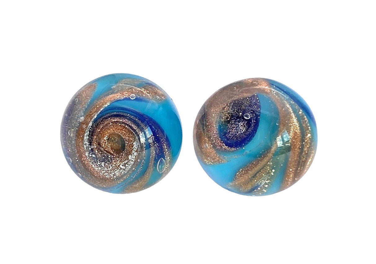 Two round marbles with swirling patterns of blue, gold, and silver hues create a mesmerizing design. Handmade in Murano, these marbles are set against a plain white background, highlighting their intricate details, reminiscent of the artistry found in Murano Glass Stud Earrings with Gold Aventurine Swirls by Murano Passion.