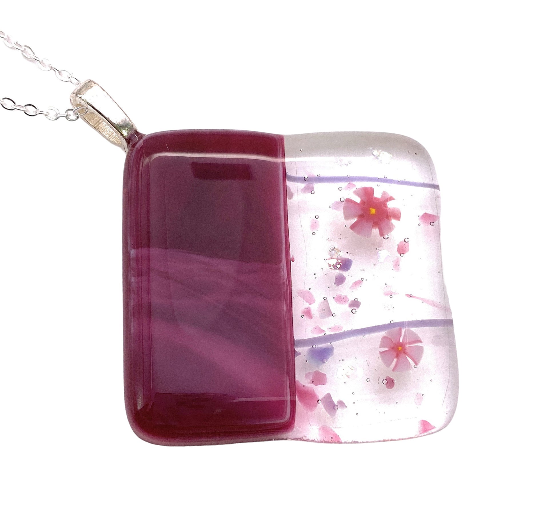 This Handmade Dichroic Glass Pendant by Calon Glass features a chunky design measuring 4cm x 4cm, combined with a silver chain. It boasts an eye-catching pattern consisting of a maroon section and a clear segment adorned with pink flowers and vibrant Millefiori glass fragments.