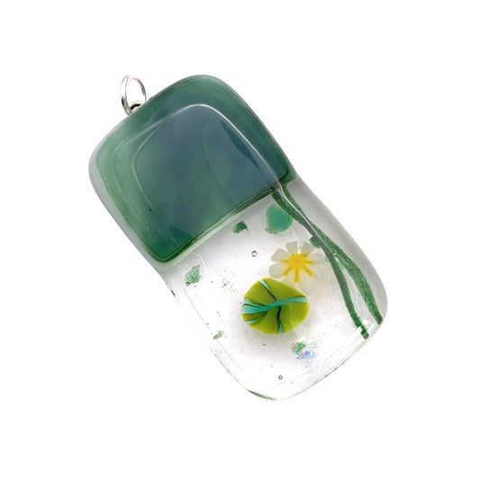 This rectangular pendant from Calon Glass, measuring 4cm x 2cm, showcases a handmade design with dichroic glass. Its top features a green color transitioning to a clear bottom, and it includes millefiori glass elements that create the appearance of a yellow flower with green accents for an organic look.