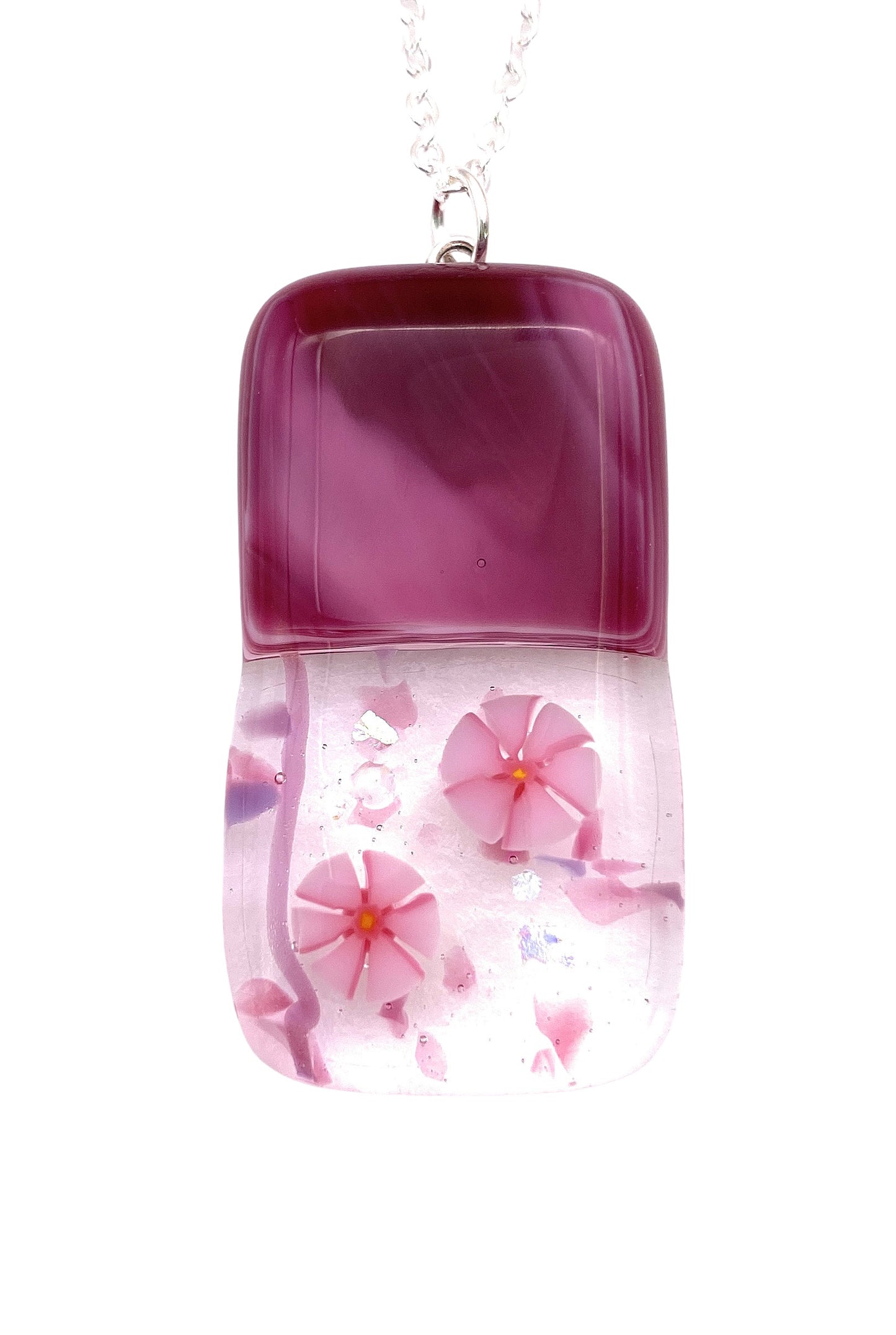 A pendant by Calon Glass made with handmade dichroic glass showcases a captivating purple gradient top and clear bottom, featuring two intricate pink millefiori flower designs embedded within. This exquisite piece measures 4cm x 2cm and hangs elegantly from a slender silver chain.